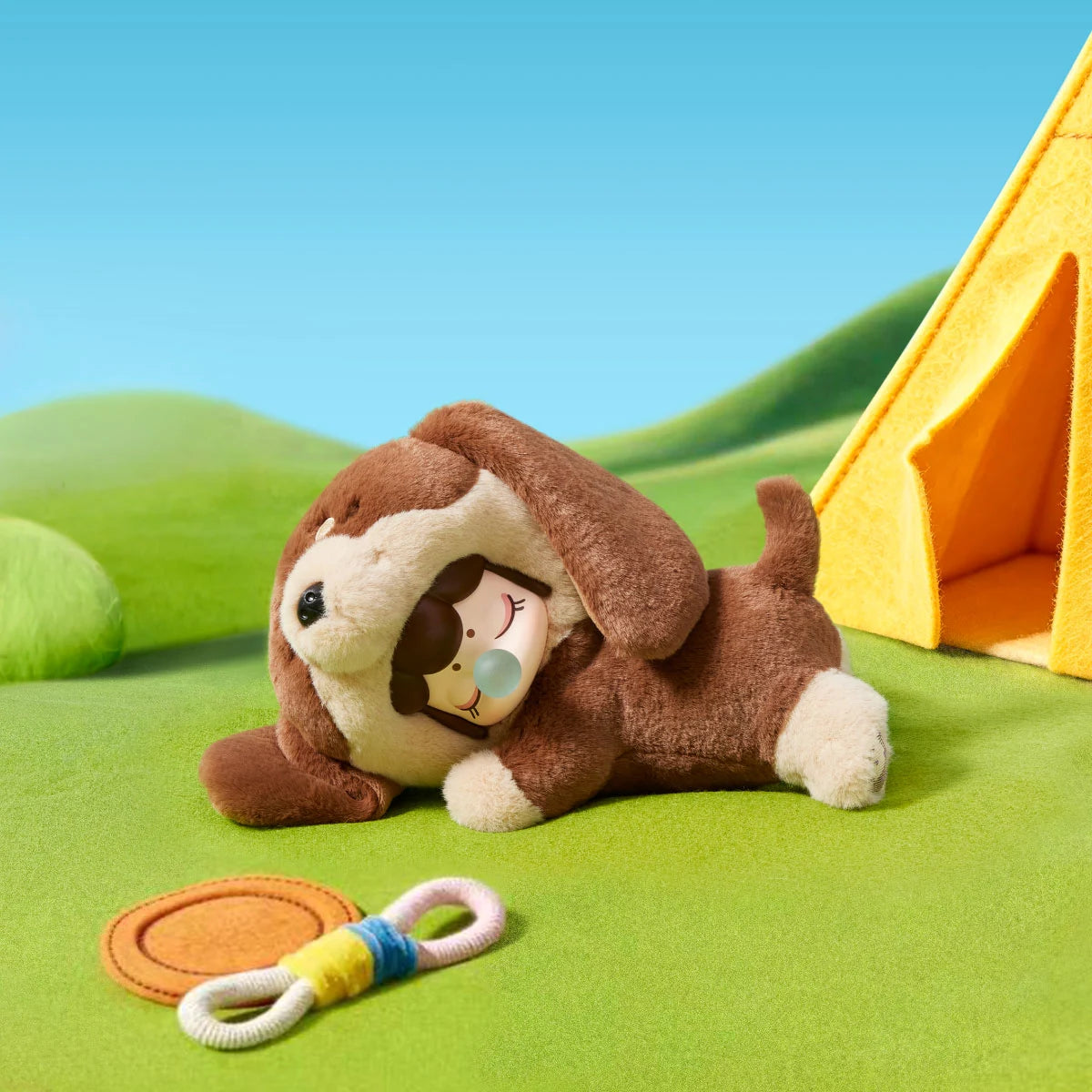 Brown-and-cream Nanci Puppy Club Dachshund plush doll lying on green felt grass beside a yellow tent, orange flying disc and braided rope toy, with floppy ears and a sleeping face.