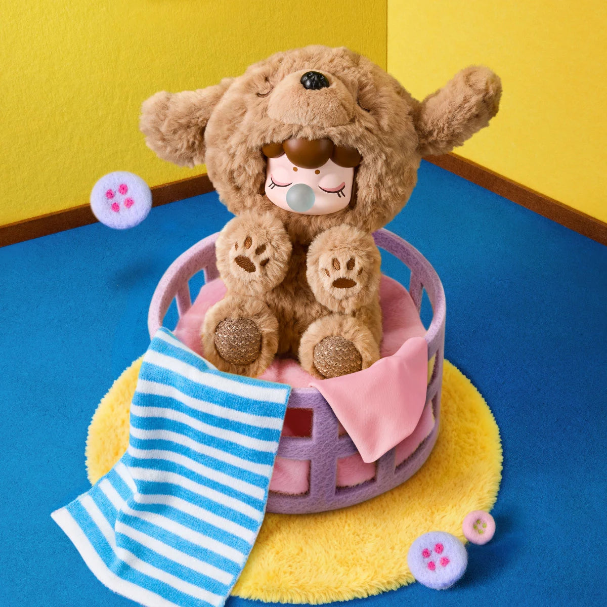 Curly brown Nanci Puppy Club Teddy plush doll sitting in a lavender basket on a yellow rug, with raised paws, embroidered paw pads, a striped towel and pink cloth.